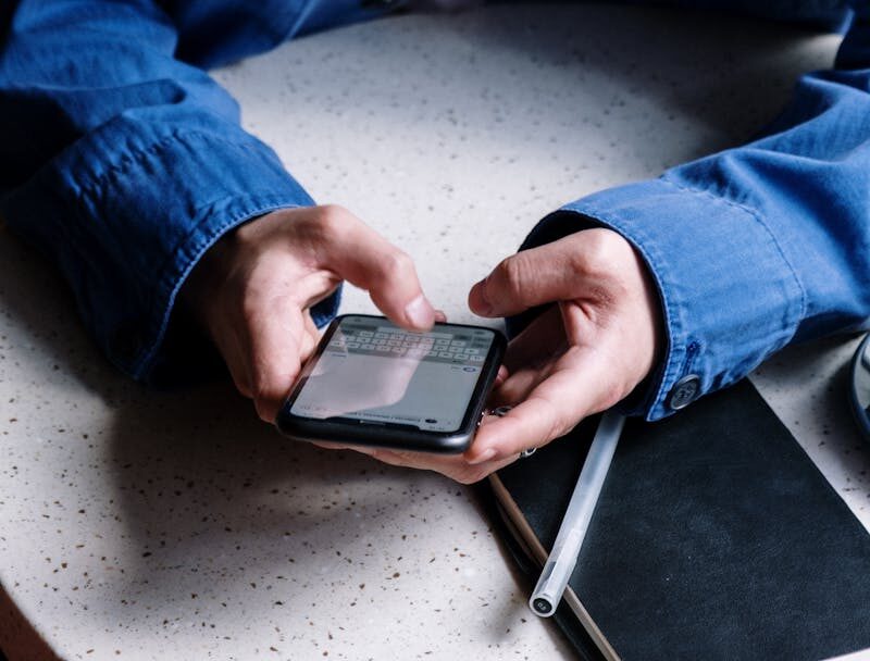 A young man in a blue shirt uses his smartphone in a cozy café, focusing on typing messages.